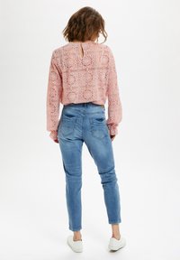 Blusa rosa in pizzo con maniche lunghe e schiena a oblò, abbinata a jeans skinny in denim azzurro chiaro e sneakers bianche.