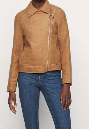 Persoon draagt een tankleurige, wollen jas met een asymmetrische ritssluiting aan de voorkant en een ringtrekker, gecombineerd met blauw skinny jeans in mid-rise.