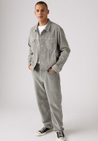 Man staat met zijn handen in zijn zakken, gekleed in een lichtgrijs corduroy jasje en broek, een wit t-shirt en zwarte low-top sneakers, tegen een effen achtergrond.