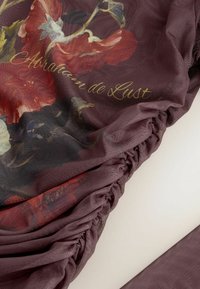 Tissu bordeaux transparent aux bords froncés, présentant un grand motif floral rouge et noir avec un texte doré en cursive « Amour de Lust ».