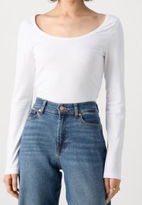 Vit långärmad topp med en bred rund halsringning, kombinerad med högmidjade blå denimjeans som har subtila blekningar och en midjebandknapp.