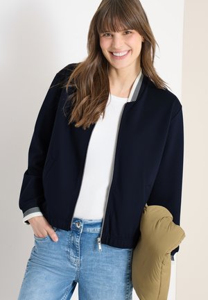 Donna sorridente con lunghi capelli castani che indossa una giacca bomber blu navy sopra una maglietta bianca e jeans azzurri, tiene una giacca imbottita beige al braccio.