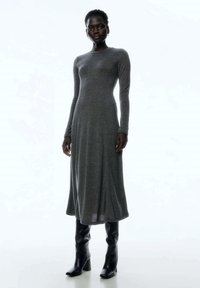 Langärmliges graues Kleid aus Strickstoff, oben figurbetont und am unteren Ende ausgestellt. Kombiniert mit knielangen schwarzen Stiefeln mit Absatz.