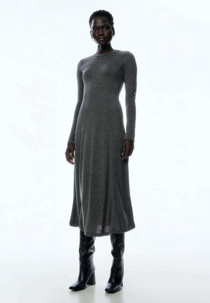 Langärmliges graues Kleid aus Strickstoff, oben figurbetont und am unteren Ende ausgestellt. Kombiniert mit knielangen schwarzen Stiefeln mit Absatz.