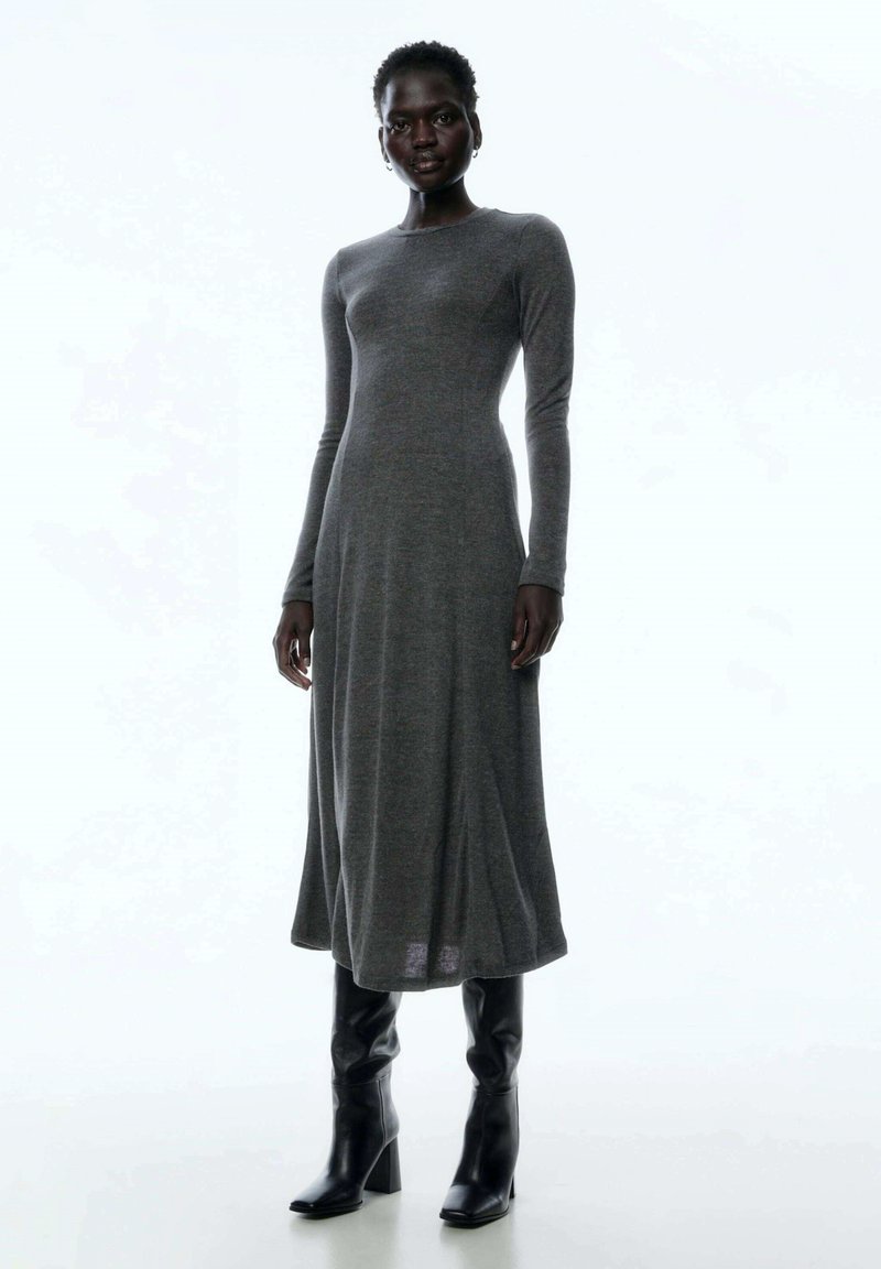 Langärmliges graues Kleid aus Strickstoff, oben figurbetont und am unteren Ende ausgestellt. Kombiniert mit knielangen schwarzen Stiefeln mit Absatz.