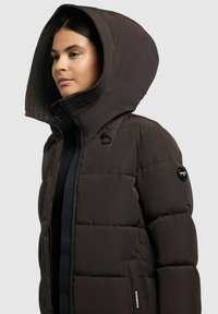 Doudoune marron avec une grande capuche, texture matelassée, fermeture éclair à l'avant et poches latérales. Présente un logo sur la manche et un tissu robuste.