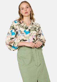 Blusa floral con grandes patrones de flores azules y rosas sobre un fondo claro, con mangas abullonadas y botones en la parte delantera. Combinada con una falda verde.