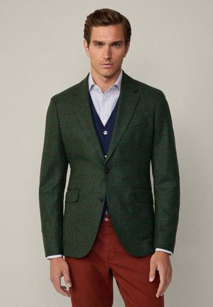 Dunkelgrüner Tweed-Blazer mit dezentem Karomuster, Reverskragen, zwei Fronttaschen und einreihigem Knopfverschluss, darüber getragen eine marineblaue Weste.