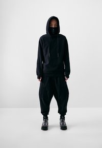 Schwarzer Kapuzenpullover mit Kordeln, passende lockere Hose und gemusterte schwarz-graue Schuhe. Glattes Material ohne sichtbare Logos.