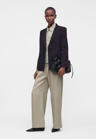 Femme portant un blazer noir à double boutonnage, une chemise rayée boutonnée, un pantalon beige à jambes larges, des chaussures noires pointues, et tenant un sac à main noir.