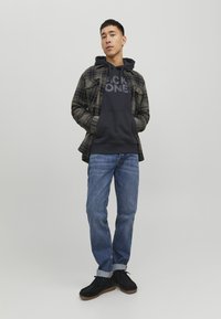 Camicia a quadri verde scuro e grigia sovrapposta a una felpa blu navy con testo grande; jeans blu e scarpe nere. Design semplice e vestibilità rilassata.