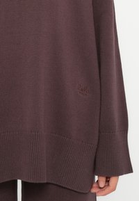 Close-up da mão de uma pessoa ao lado de um suéter de malha em mauve escuro, com nervuras soltas, e um pequeno logotipo bordado de lado.