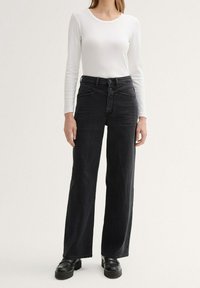 Jeans negros de pierna ancha, confeccionados en denim, con una cintura alta, dos bolsillos frontales, cierre de botón y una textura sutil. Combinados con una blusa blanca de manga larga.