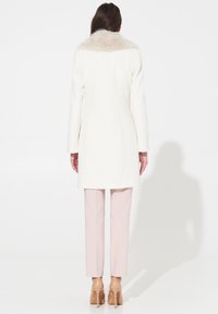 Cappotto bianco con colletto di pelliccia, sagomatura impeccabile, maniche lunghe e texture liscia. Abbinato a pantaloni slim rosa chiaro e tacchi beige.