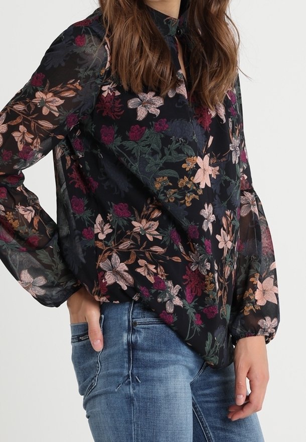 Femme portant un chemisier noir transparent à manches longues avec un motif floral multicolore et un jean bleu, une main dans la poche.