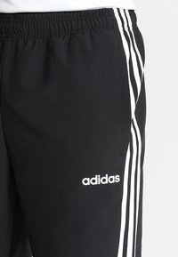 Černé adidas teplákové kalhoty s elastickým pasem, třemi bílými bočními pruhy a logem adidas na horní části stehna.