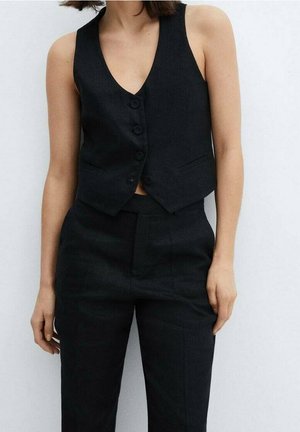 Person trägt eine schwarze ärmellose, geknöpfte Weste und passende schwarze, hoch taillierte Hose vor einem einfarbig hellen Hintergrund.