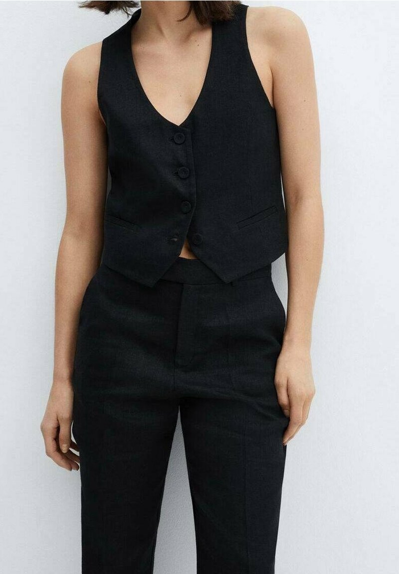Personne portant un gilet noir sans manches boutonné et un pantalon noir taille haute assorti sur un fond clair uni.