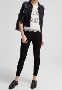 Chaqueta de mezclilla negra sobre una blusa de encaje blanca con dobladillo en picos, combinada con jeans ajustados negros y zapatos negros con cordones.