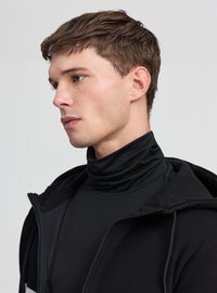 Felpa nera con zip e tessuto morbido, indossata sopra un dolcevita nero abbinato. Presenta un design aderente e un colletto alto. Stile minimalista.
