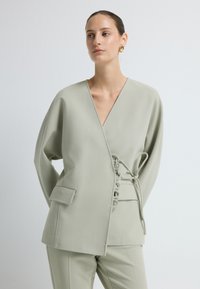 Jasnozielona kurtka o luźnym kroju z głębokim dekoltem w serek, szerokimi rękawami i detalem w postaci wiązania po bokach. Teksturowany materiał z kieszenią po lewej stronie.