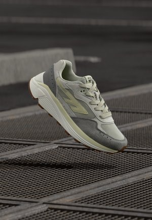 Pilkšvos ir smėlio spalvos sportbačiai, pakibę virš metalinės grotelės miesto lauko aplinkoje, demonstruoja tekstūruotą padą ir raišteliais sujungtą viršų.