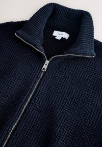 Veste en maille texturée bleu marine avec une fermeture éclair argentée sur le devant et un col montant côtelé, l'étiquette indique "NEXT" à l'intérieur du col.
