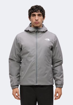 Voksen mand iført en grå hættetrøje med lynlås, elastiske manchetter og The North Face-logo på brystet, stående foran en ensfarvet baggrund.