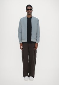 Pelēks bomber jakas ar priekšējo rāvējslēdzēju, uzvilkts virs melnas krekla, apvienots ar brūnām cargo biksēm un baltām kedām. Modelis stāv uz balta fona.