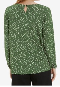 Femme portant un chemisier vert à manches longues avec un motif floral blanc et une petite ouverture en forme de larme au niveau de l'encolure dos.