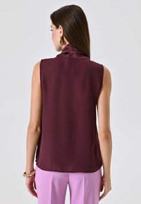 Top senza maniche color marrone scuro con collo alto, realizzato in tessuto liscio, visto da dietro, abbinato a pantaloni viola chiaro.