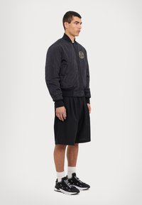 Svart bomberjacka med dragkedja framtill, elastiska muddar och sidofickor. Har en guld logotypapplikation på bröstet. Bärs med svarta shorts och sneakers.