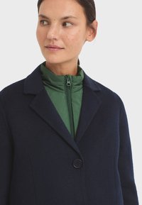 Abrigo de lana azul marino con cierre de botones y solapas con muesca, superpuesto sobre una chaqueta verde con cremallera. Textura suave y corte entallado.