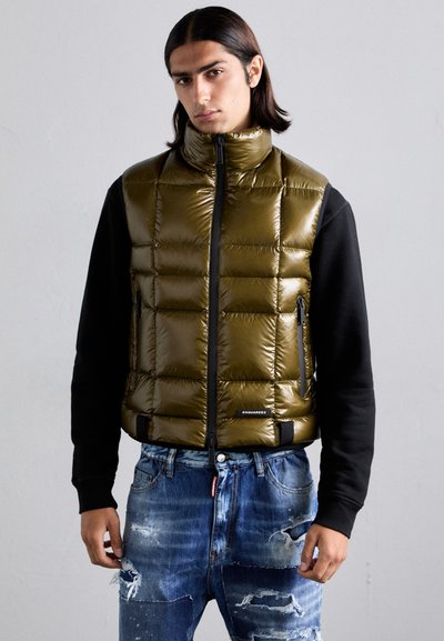 Gilet rembourré vert olive avec col montant, fermeture zip, poches latérales et manches noires, associé à un jean bleu déchiré.