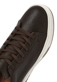 Sneaker de cuero marrón con superficie texturizada, punta redonda y cordones marrones oscuros. Presenta una suela de goma blanca y detalle de costura lateral.