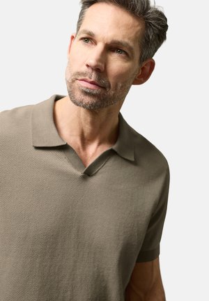 Mann mit salt-and-pepper Haaren, der ein strukturiertes, olivgrünes Polohemd mit kurzen Ärmeln trägt und leicht zur Seite blickt vor einem schlichten Hintergrund.