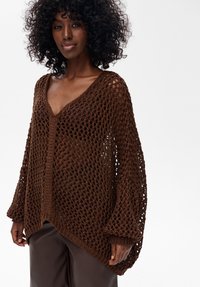 Brązowy sweter robiony na drutach o luźnym, otwartym kroju, z dekoltem w serek i długimi rękawami. Teksturalny splot dodaje wizualnego zainteresowania i głębi.