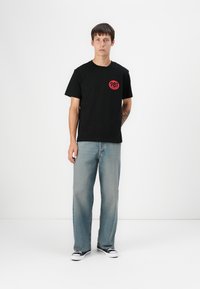 Tricou negru din bumbac, cu mâneci scurte și un logo circular roșu cu "981" pe pieptul stâng, asortat cu blugi drepți de un albastru estompat.