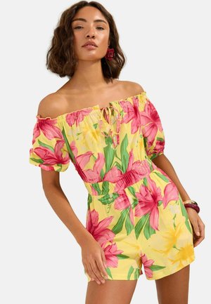 Frau trägt gelben schulterfreien Overall mit pinkgrünem Blumenmuster, kurzen Ärmeln und elastischer Taille, accessoriert mit Blumenohrring und Armbändern.