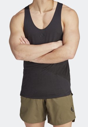 Homme aux bras croisés portant un débardeur noir sans manches et un short Adidas vert olive, sur fond neutre.