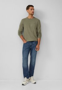 Olivgrünes Langarmhemd, blaue Jeans, hellfarbene Sneakers mit strukturierten Akzenten. Das Modell steht lässig mit einer Hand in der Tasche.