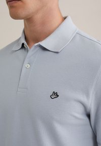 Polo de culoare albastră deschisă, cu un material texturat, trei butoane și un mic logo brodat cu o pasăre neagră pe partea stângă a pieptului.