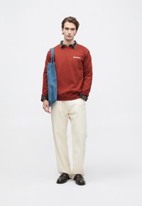 Νεαρός άνδρας με καμένου πορτοκαλί φούτερ Barbour, κρεμ παντελόνι, μαύρα παπούτσια και κρατώντας μια μπλε τσάντα ώμου, στέκεται χαλαρός.