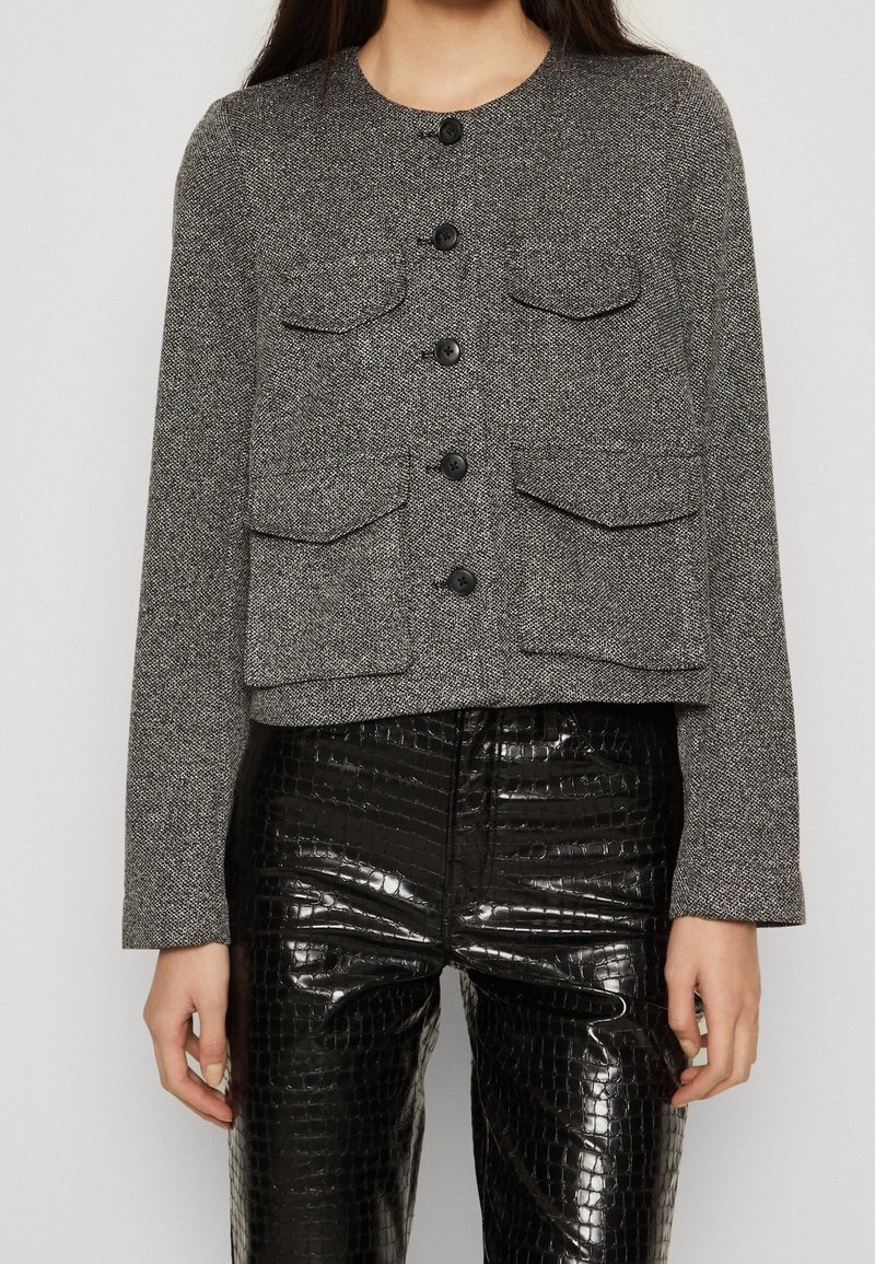Femme portant une veste grise boutonnée avec quatre poches à rabat et un pantalon en similicuir noir brillant texturé.