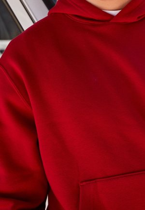 Person trägt einen leuchtend roten Hoodie mit Fronttasche und weißem Unterhemd, teilweise Ansicht vom Brustkorb bis zum Kinn.