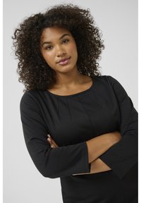 Haut noir à manches longues avec un col rond et des plis décoratifs sur la poitrine. Il présente un tissu lisse et élastique ainsi qu'une coupe ajustée.