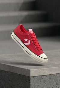 Baskets en toile rouge avec semelle en caoutchouc blanche, arborant un logo étoile blanc et des accents roses. Conception classique à lacets avec un bout arrondi.