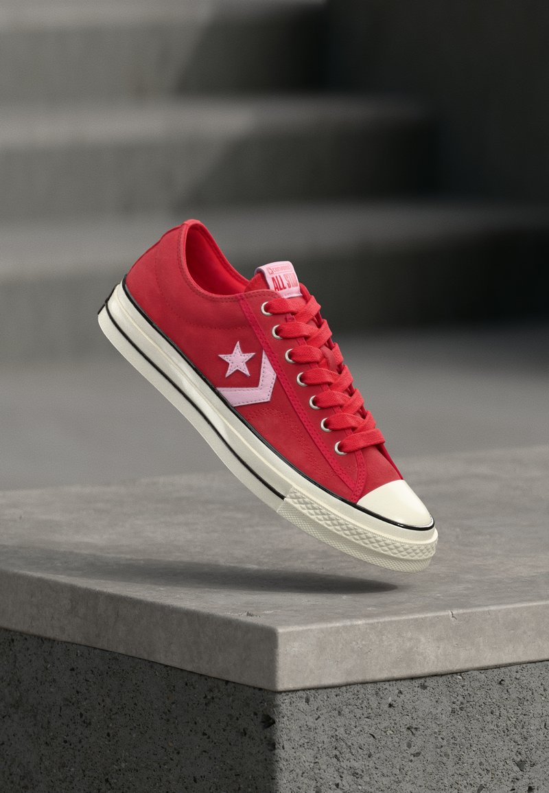 Baskets en toile rouge avec semelle en caoutchouc blanche, arborant un logo étoile blanc et des accents roses. Conception classique à lacets avec un bout arrondi.