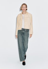 Donna che cammina indossando una giacca di camoscio beige, una maglietta grigia chiara, jeans blu larghi e scarpe nere su uno sfondo bianco semplice.