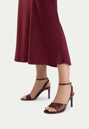 Pies con sandalias de tacón alto color burdeos con tiras y hebillas en el tobillo, combinadas con una falda midi burdeos.